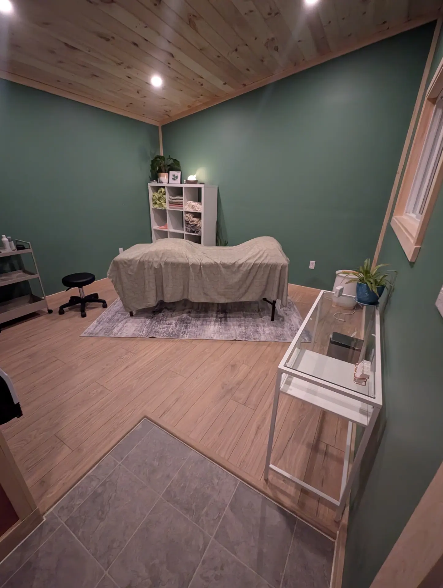 Haley’s treatment room in Hebbville, Nova Scotia: a draped massage table on an area rug, sage green walls, light wood flooring and a pine plank ceiling